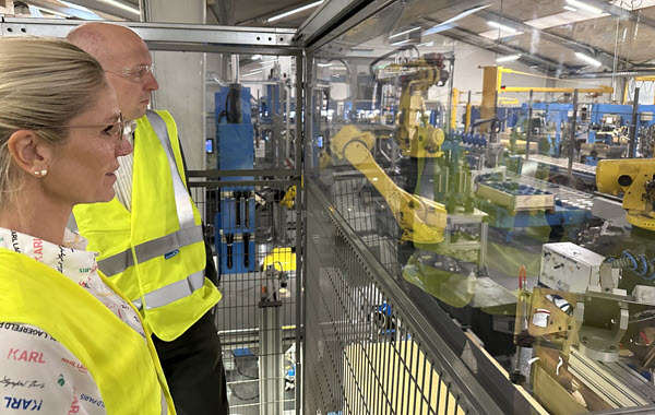 To personer i gule veste ser ind i et område med robotter og produktionsudstyr bag glas.